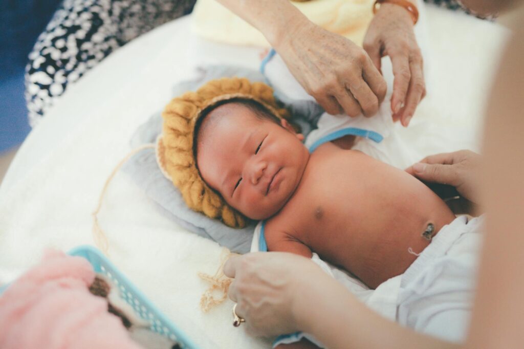 A calm newborn baby lying on a bed with caregivers adjusting a blanket indoors, captured in soft light.