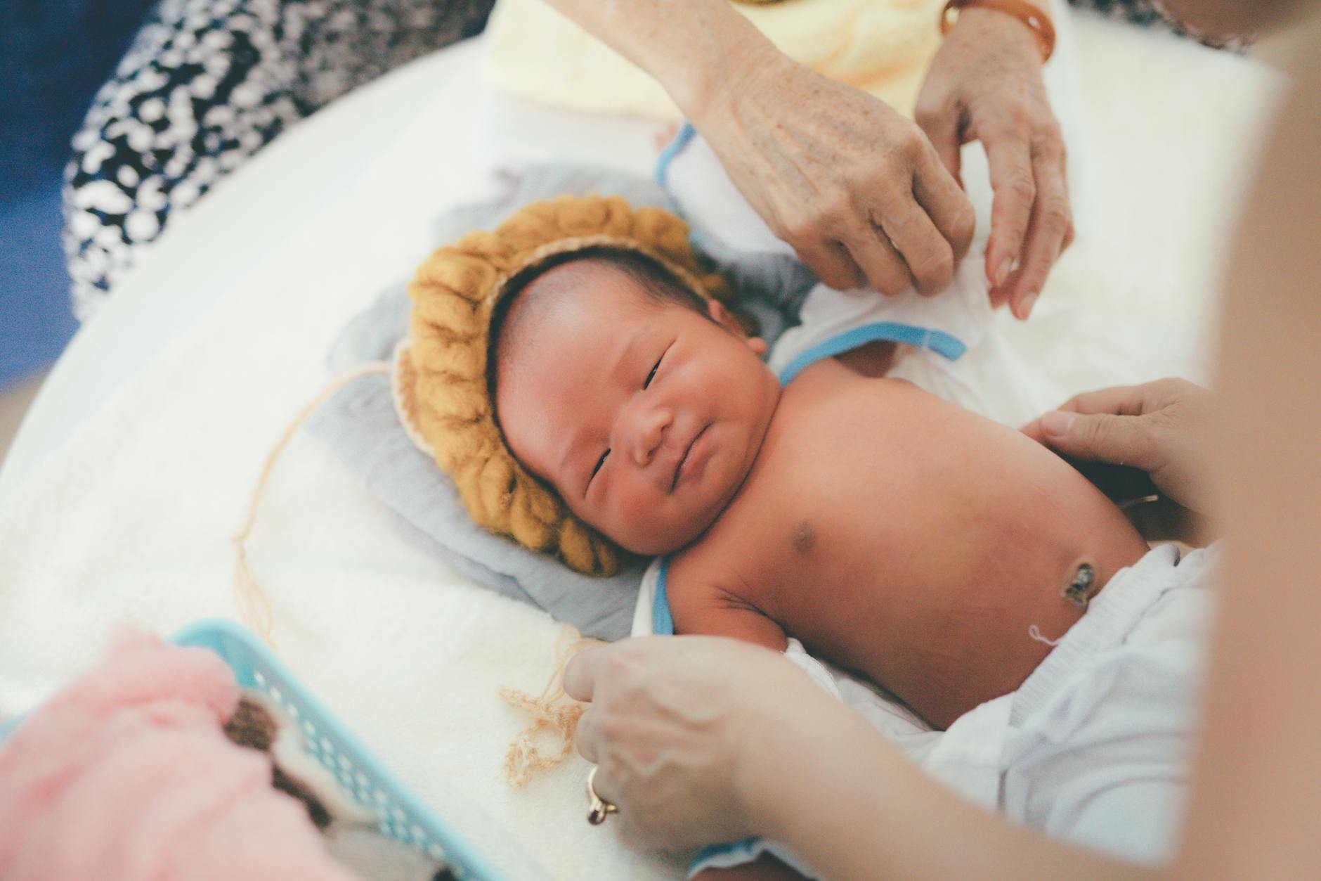 pexels-photo-9300617-9300617 A calm newborn baby lying on a bed with caregivers adjusting a blanket indoors, captured in soft light.
